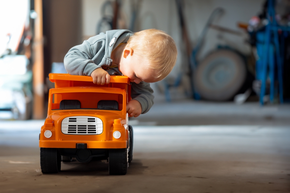 A toddler leaning over a large orange toy dump truck in a garage, used to illustrate the potential risks and injuries associated with defective toys.