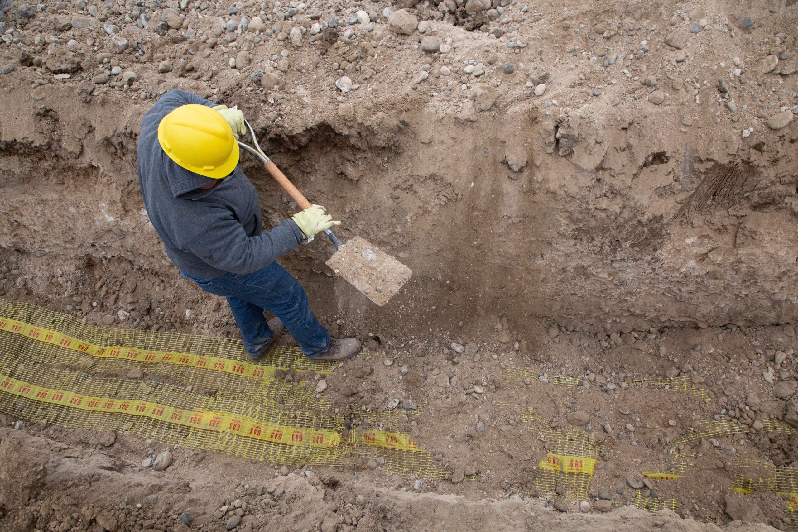 Aerial view of a construction worker in a yellow hard hat and blue work clothes digging in an excavation trench, with yellow gas line warning tape visible across the trench floor, illustrating trench safety hazards on New Jersey construction sites.