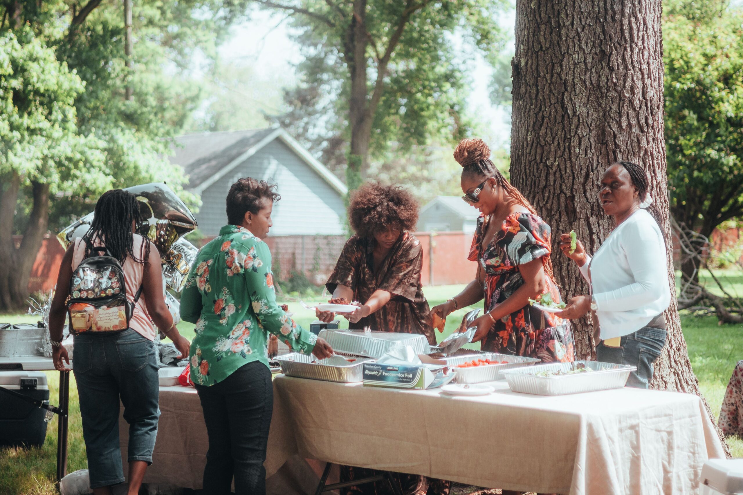 Women serving food at an outdoor backyard graduation party, gathered around a buffet table with aluminum catering trays under a large tree, with green grass and a house visible in the background
