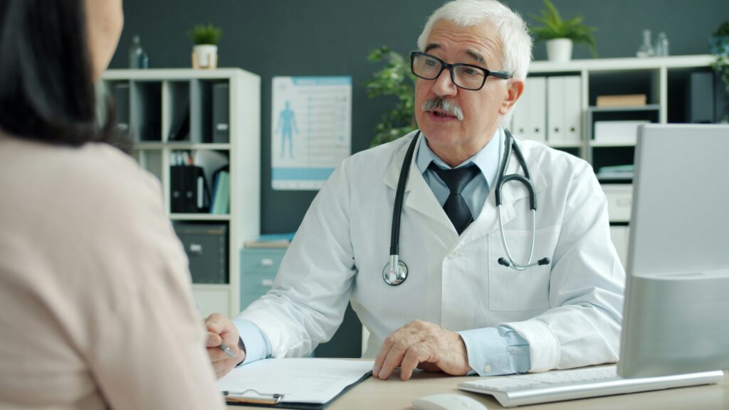 Older doctor talking with a female patient at his desk during an office visit, showing how medical gaslighting can happen in an appointment.